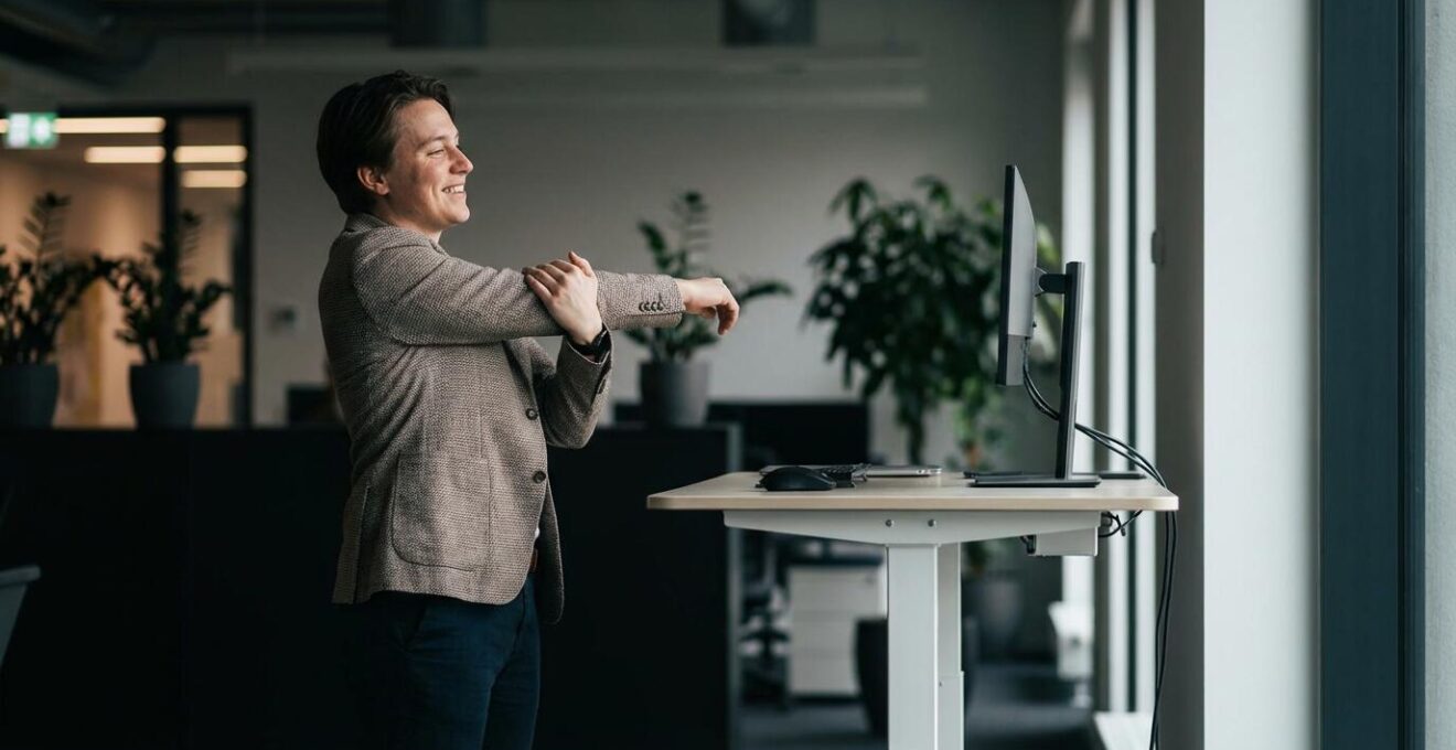 Business professional performing a standing desk stretch routine in a modern office with natural lighting